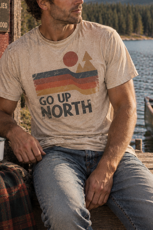 A man sitting on a wooden dock by a lake wearing a beige graphic t-shirt with a retro sunset design and the text 'GO UP NORTH'.